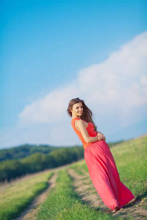 Portrait of barefoot woman in long red dress walking on the road in the green field in summerの写真素材