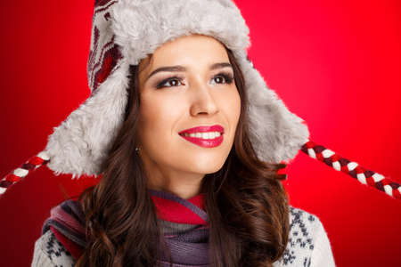 Portrait of beautiful woman in warm clothing and winter hat with bright makeup on red background ...