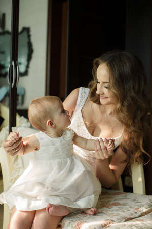 Mother with little baby in home interior young beautiful mom hugging daughter, pretty brunet woman playing with cute small girl, smiling girl holding adorable sweet child, happy family conceptの写真素材
