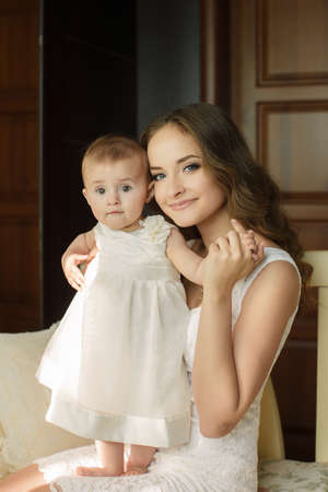Mother with little baby in home interior young beautiful mom hugging daughter, pretty brunet woman playing with cute small girl, smiling girl holding adorable sweet child, happy family conceptの写真素材