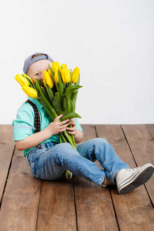 Smiling boy holding a bouquet of yellow tulips in hands sitting on wooden floor