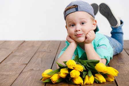 Smiling boy holding a bouquet of yellow tulips in hands sitting on wooden floorの写真素材