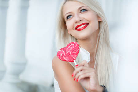 A young woman with a lollipop  Happy girl with candy in her hands  Licking candy  Outdoors lifestyle  Elegant blonde with a lollipop in the shape of a heartの写真素材