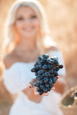 Smiling woman with basket of grapes sitting in summer sunny field. Beautiful blonde woman in straw hat with harvest outdoor.の写真素材