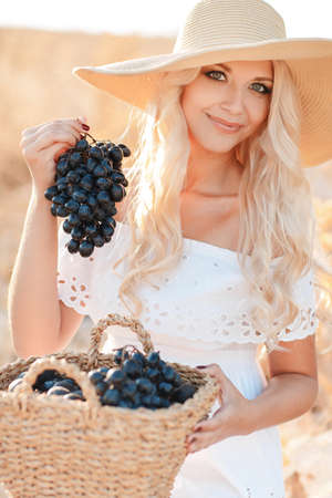 Smiling woman with basket of grapes sitting in summer sunny field. Beautiful blonde woman in straw hat with harvest outdoor.の写真素材