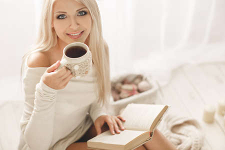 Soft portrait of a blonde with gray eyes, drinking hot coffee and reading a book at home. Young beautiful woman in warm knitted clothes handmade home. Fall, winter season.の写真素材