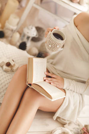 Soft portrait of a blonde with gray eyes, drinking hot coffee and reading a book at home. Young beautiful woman in warm knitted clothes handmade home. Fall, winter season.の写真素材