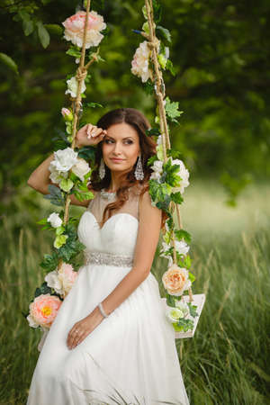 Portrait of the beautiful bride in the white wedding dress sitting on a swing in the open air in green park in the summerの写真素材