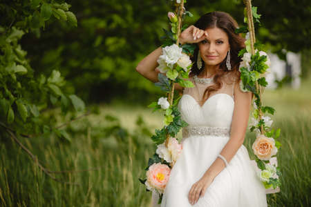 Portrait of the beautiful bride in the white wedding dress sitting on a swing in the open air in green park in the summerの写真素材