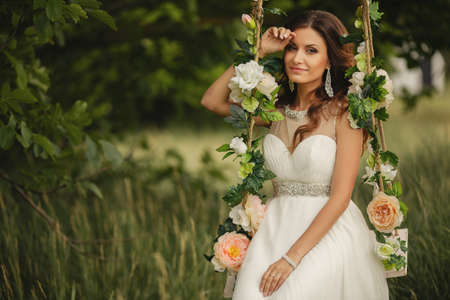 Portrait of the beautiful bride in the white wedding dress sitting on a swing in the open air in green park in the summerの写真素材