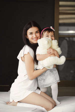 Portrait plays a mother with a small daughter. A beautiful woman is a brunette in a white nightgown, with long straight hair and gray - green eyes, playing with her little daughter and a white Teddy bear in the room on the bed.の写真素材