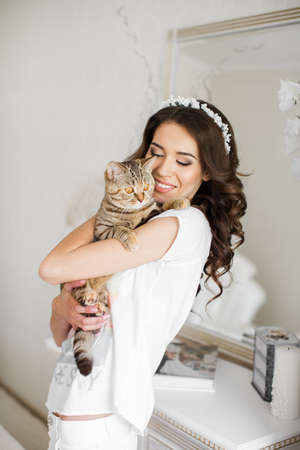 A young bride, brunette , long curly hair, wedding wreath on his head, in the night white shirt , holding a gray cat, preparing for the wedding night in front of the mirror in a beautiful white bedroom.の写真素材
