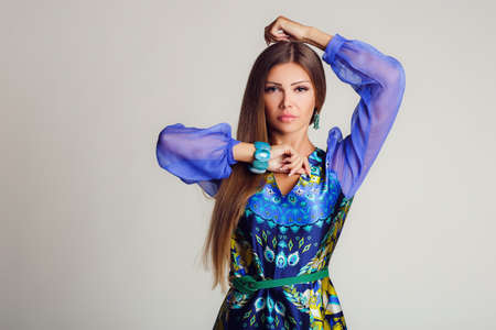 Portrait-style - professional model on a light background.A young woman, a brunette with long straight hair, with gray - green eyes, light makeup, a mole on his left cheek,makes the display bright coloring blue dress with transparent sleeves, beige high hの写真素材