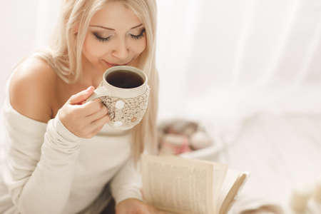 Young beautiful woman - blonde, with light make-up, wearing a light - beige knitted, short dress, sitting on a white bed with a morning cup of hot coffee in his hands.の写真素材