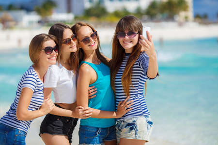 Beautiful girls are photographed on a tropical resort on the background of the beach and ocean.Four young beautiful girls - brunette, long straight hair, in shorts and T-shirts, sun glasses, with a beautiful smile, photographed on a smartphone on the backの写真素材