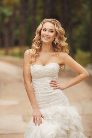 Bride in wedding dress posing for a photograph in the Park in the summer. Very beautiful woman in wedding white dress, with long, curly, blond hair, blue eyes, beautiful smile and beautiful earrings, posing in summer green park.の写真素材