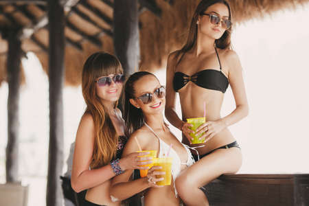 three beautiful sexy women in bikini in bar on Hawaii at beach bar. Beautiful girls enjoying alcoholic beverage cocktail outside. Smiling happy Caucasian women on Hawaiian beach.の写真素材