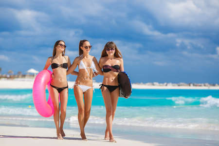 Three slim young girls in bikinis on the beach. Group of Three Beautiful Attractive Young Women Walking on the Beach. happy girl friends having fun at the beach while on vacationの写真素材