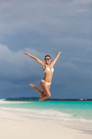 A beautiful woman, a brunette with pinned up on the back of the head hair,in a white bikini, sunglasses , having fun on the white sandy tropical beach, jumping high in the blue ocean.の写真素材