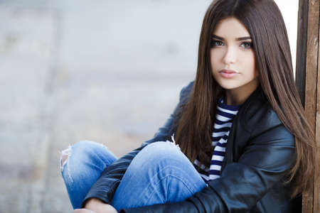 Beautiful young girl, Caucasian appearance, with dark, long, straight hair, brown eyes and beautiful dark eyebrows, wearing a striped shirt, blue jeans and black leather jacket, sitting on the street on the sidewalkの写真素材