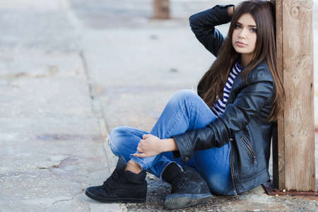 Beautiful young girl, Caucasian appearance, with dark, long, straight hair, brown eyes and beautiful dark eyebrows, wearing a striped shirt, blue jeans and black leather jacket, sitting on the street on the sidewalkの写真素材