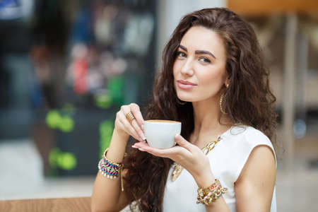 Young brunette with long curly hair and brown eyes, wearing gold jewelry, bracelet, earrings, rings, necklaces, dressed in white  blouse, standing near a brown table in a cafe, drinking coffee from a white cup.の写真素材