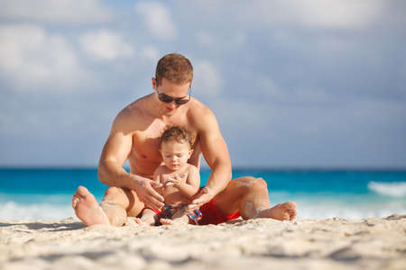 Handsome muscular man of Asian appearance with short hair,dressed in red shorts,plays on a tropical beach with her young son with curly hair, wearing plaid shorts,near blue ocean.の写真素材
