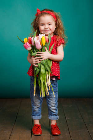 Studio portrait of a pretty little girl child with long curly hair and brown eyes with a large bouquet of tulips in hand, dressed in a red dress with white polka dots, red bandage on his head and blue jeans, isolated on a blue background.の写真素材