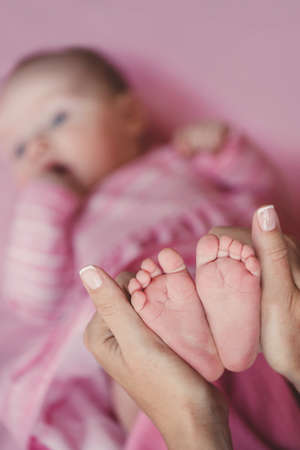 Newborn baby girl wearing a pink dress, lying on his back in his bed on a blanket pink color, bare little feet daughter tenderly holds the young mother, raising the leg over the blanket.の写真素材