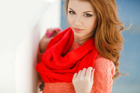 Portrait of a beautiful young woman with long hair chestnut, hazel eyes and light make-up, wearing a pink knitted jacket and red scarf around his neck, a beautiful smile and straight white teeth, posing for a photograph near the white building.の写真素材