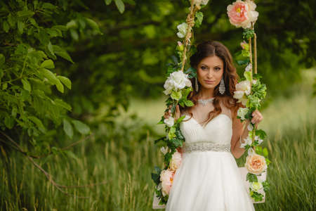 Young beautiful bride in a white wedding dress swinging on a swing decorated with roses in a green Park in the summer, earrings in your ears,on your left hand bracelet,brown eyes and long curly hair,wearing your wedding ring.の写真素材