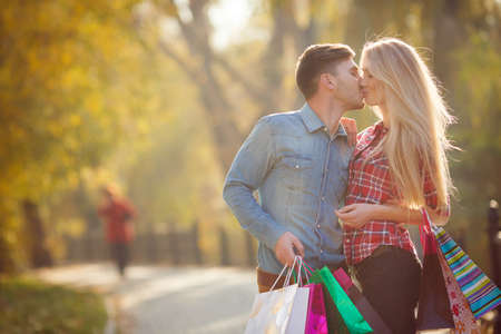 Young couple in love, boy-short-haired brunette in a blue denim shirt and brown trousers and a girl - blonde with long hair, dressed in a red plaid shirt, posing in autumn park with paper bags for shopping.の写真素材