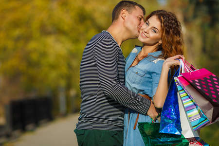 Happy young family - a man,a brunette with short hair in a striped shirt and green pants and woman-brunette with long curly hair wearing a blue denim dress with a brown belt,go shopping to the Mall through a city Park with paper bagsの写真素材