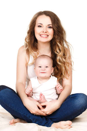Portrait of a mother and baby isolated on white background on a light beige blanket,brown-eyed baby with a fluffy short hair,in a white t-shirt and the mother is a brunette with long curly hair and grey eyes,wearing a white t-shirt,smiling and posingの写真素材