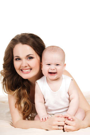 Portrait of a mother and baby isolated on white background on a light beige blanket,brown-eyed baby with a fluffy short hair,in a white t-shirt and the mother is a brunette with long curly hair and grey eyes,wearing a white t-shirt,smiling and posingの写真素材