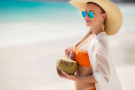 A woman with a beautiful figure, brunette with long straight hair in orange bikini, in a large straw hat with a light transparent white scarf draped behind dark glasses standing on a beach, drinking through a straw coconut milk.の写真素材