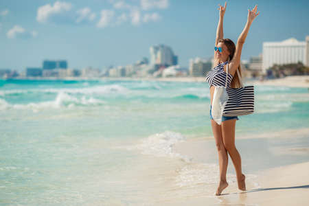 A beautiful young woman,a brunette with long straight hair,in blue shorts, wearing sun glasses with blue glasses, striped beach bag and a striped t-shirt,standing near the ocean enjoying the vacation on a tropical resort.の写真素材