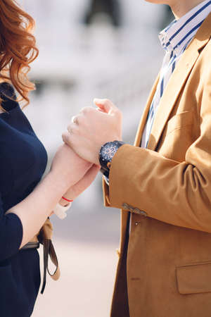 A young,stylish couple in love,a woman in a short dark-colored dress and a man in a light brown jacket and white in a dark striped shirt,standing next to each other on a city street and holding handsの写真素材