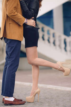 Young man in a dark blue jeans, brown shoes and white socks and a woman in beige and dark shoes short dress, kissing on the street in the city on the background of a white building with a handrail, photographed the legs of a young couple.の写真素材