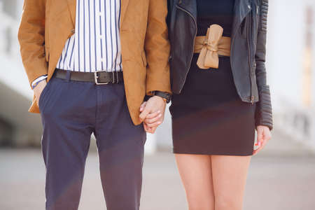 A young,stylish couple in love,a woman in a short dark-colored dress and a man in a light brown jacket and white in a dark striped shirt,standing next to each other on a city street and holding handsの写真素材