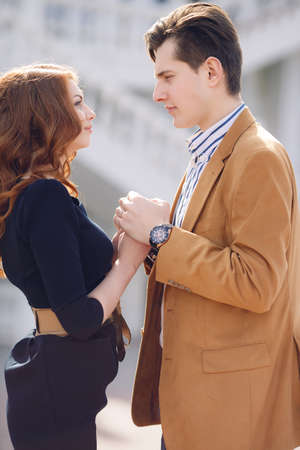 Happy couple in love,a man with dark hair in a light brown jacket and white striped shirt and a beautiful woman with long red curly hair,in a black dress,gently hugging and talking standing on the street spring cityの写真素材