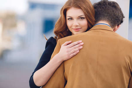 Happy couple in love,a man with dark hair in a light brown jacket and white striped shirt and a beautiful woman with long red curly hair,in a black dress,gently hugging and talking standing on the street spring cityの写真素材