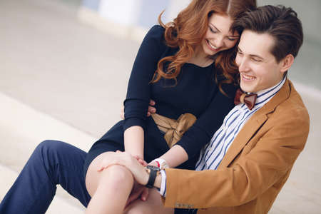 Happy couple in love, man with dark hair in a light brown coat and white striped shirt and a beautiful woman with long curly hair red color in a black dress, sitting on the lap of the man affectionately hugging each other outdoors spring cityの写真素材
