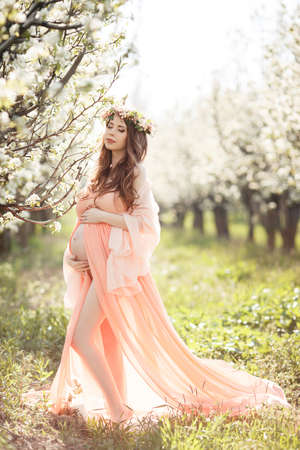 Young beautiful pregnant womanbrunette with long curly hairwears on his head a wreath of flowersdressed in a nightlong chemise pink colorpink pantiesstanding barefoot on the green grass in a lush white flowers fruit garden in the spring.の写真素材