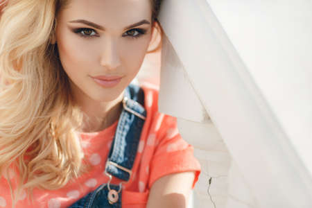 Summer portrait of a beautiful woman. Very beautiful blonde girl with big brown eyes bushy long hair dressed in a pink Tshirt with white polka dots and denim overalls in dark blue green park resting in a sunny summer dayの写真素材