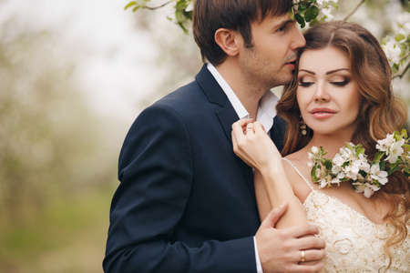 Wedding couplethe groom is a young darkhaired man in a black suit and pink bow tiebeautiful bridebrunette with long curly hair in a white wedding dress on her head a wreathposing embracing in the Park among the blooming trees.の写真素材