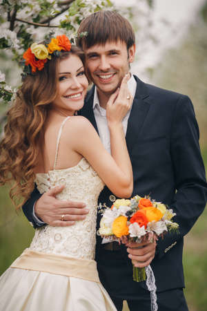 Wedding couplethe groom is a young darkhaired man in a black suit and pink bow tiebeautiful bridebrunette with long curly hair in a white wedding dress on her head a wreathposing embracing in the Park among the blooming trees.の写真素材