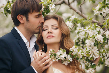 Wedding couplethe groom is a young darkhaired man in a black suit and pink bow tiebeautiful bridebrunette with long curly hair in a white wedding dress on her head a wreathposing embracing in the Park among the blooming trees.の写真素材