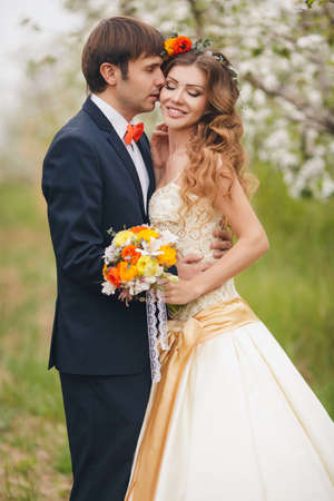 Wedding couplethe groom is a young darkhaired man in a black suit and pink bow tiebeautiful bridebrunette with long curly hair in a white wedding dress on her head a wreathposing embracing in the Park among the blooming trees.の写真素材