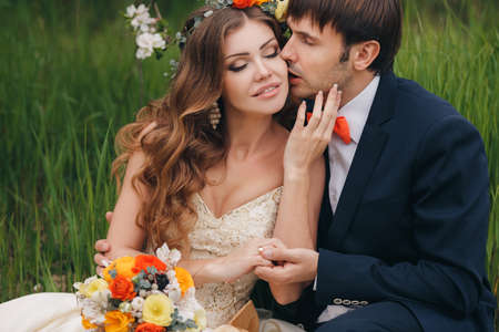 The groom is a young darkhaired man in a black suit and pink wedding tie kisses on the cheek bride beautiful brunette with long curly hair wearing a white wedding dresson her head a wreath of roses in a flowery Park in the spring.の写真素材